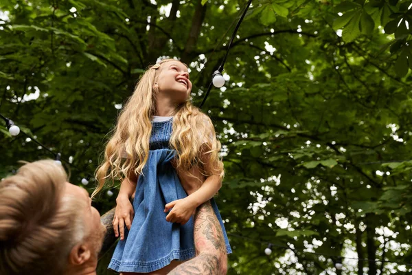 excited girl laughing in hands of tattooed father in green park, leisure and playing together