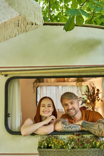 happy tattooed man and redhead woman looking out window of modern trailer home, leisure and fun