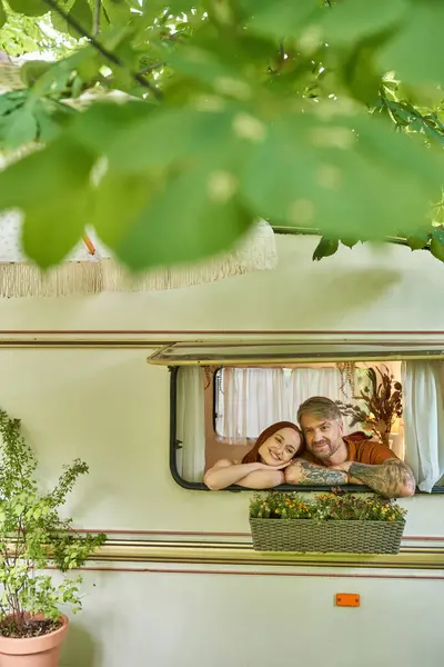 happy and stylish couple looking out window of mobile home around greenery in trailer park