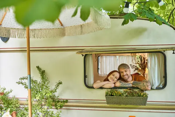 joyful and stylish couple looking out window of mobile home around greenery in trailer park