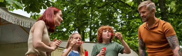 stock image joyful parents looking at cute kids blowing soap bubbles near trailer home outdoors, banner