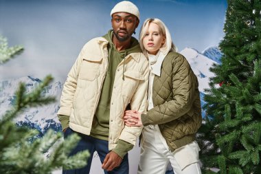 stylish multiracial couple in warm winter clothes posing together on snowy backdrop, fashion concept clipart