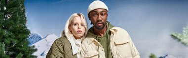 cute interracial couple in warm clothes posing together on snowy backdrop, winter fashion, banner clipart