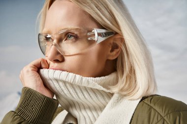 portrait of blonde woman in glasses covering mouth with her collar and looking away, winter concept clipart