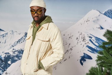 good looking man in white beanie hat with sunglasses posing next to fir tree, winter concept clipart