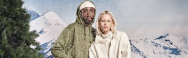 stylish multiethnic couple posing under falling snow and looking at camera, winter concept, banner clipart