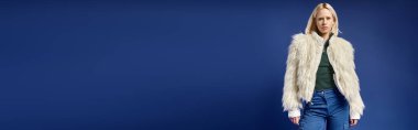 Beyaz sahte kürk ceketli, kot pantolonlu şık sarışın kadın mavi pankartta kameraya bakıyor.