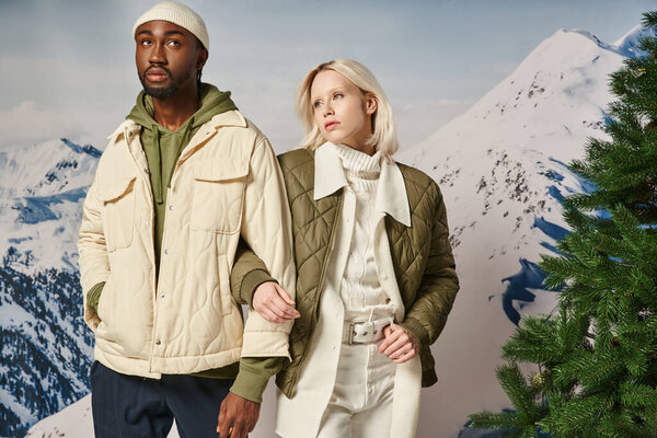 модная пара в теплых куртках рядом с сосеной позирует рука об руку, зимняя концепция