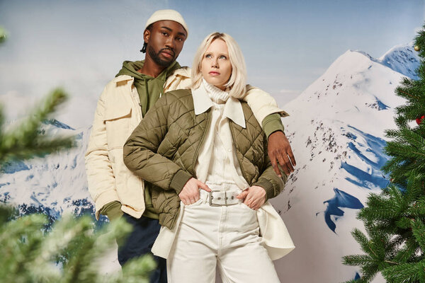 stylish diverse couple with mountain on backdrop, arm on shoulder, hands in pockets, winter concept
