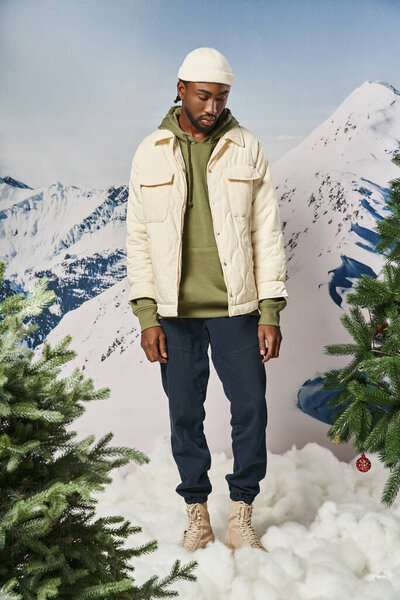 handsome african american man in beanie hat looking down with mountain backdrop, winter fashion