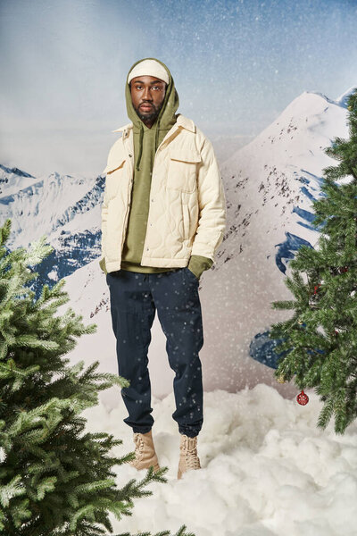 stylish good looking man posing under snowfall with hands in pockets looking at camera, winter