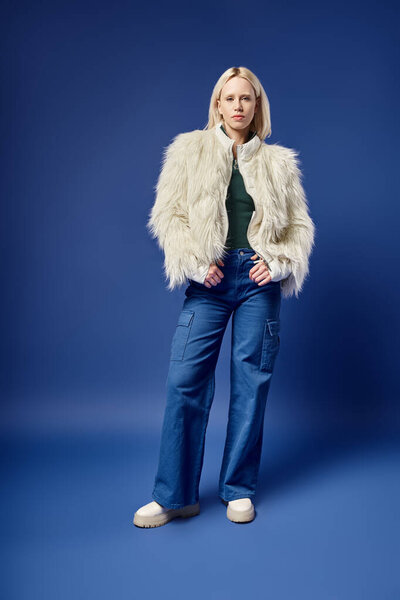 full length of stylish blonde woman in faux fur jacket and denim jeans posing on blue backdrop