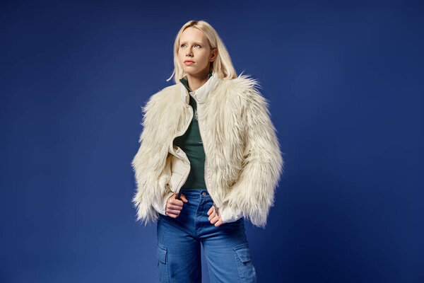 dreamy and stylish blonde woman in faux fur jacket and denim jeans posing on blue backdrop