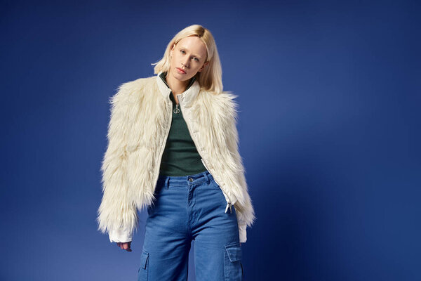 winter style, attractive young woman in faux fur jacket and denim jeans posing on blue backdrop