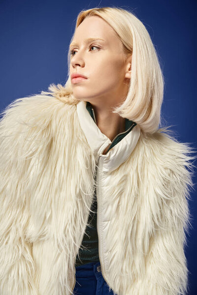 winter style, portrait of young woman in white faux fur jacket looking away on blue backdrop
