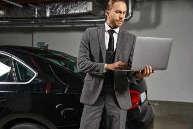 good looking man in smart suit working on laptop on parking lot near his car, business concept clipart