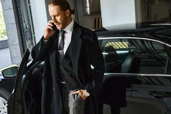 good looking elegant man in black coat talking by phone on parking lot with hand in pocket, business