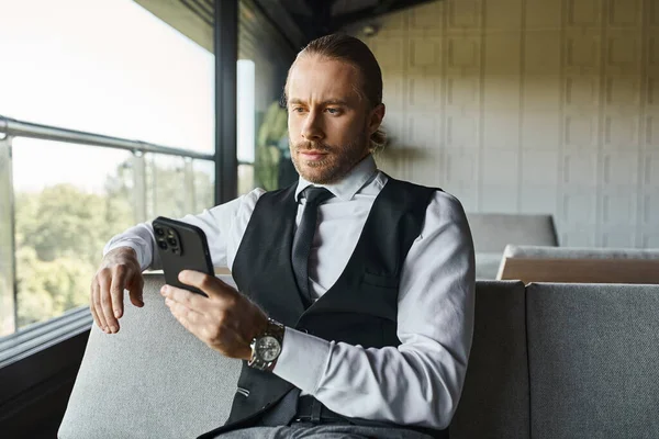 handsome red haired man in smart vest sitting on sofa and looking at mobile phone, business concept