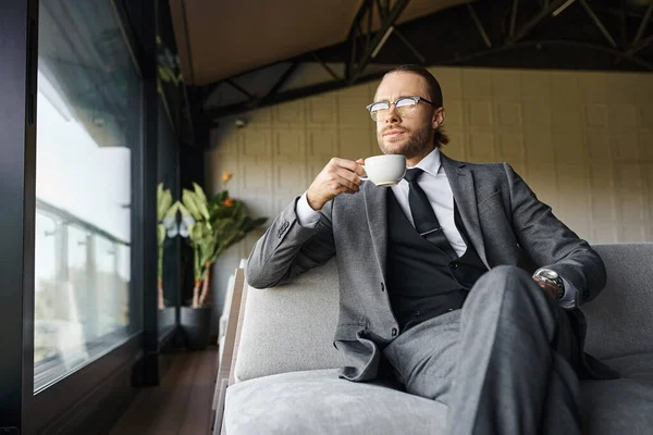 appealing man with glasses in gray suit drinking tea on sofa and looking away, business concept