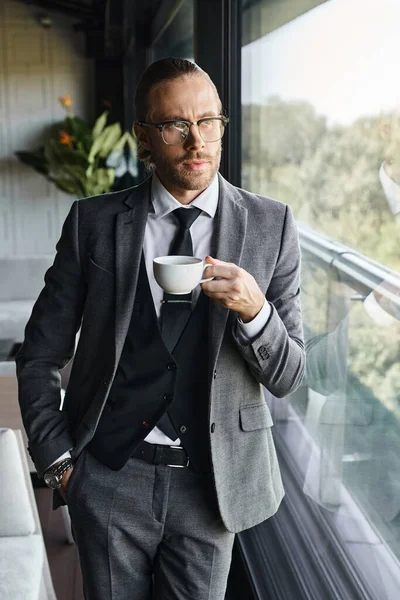 vertical shot of appealing elegant businessman in gray suit drinking tea with hand in pocket