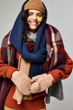 Kış tarzı, katmanlı giyinen şık bir kadın, gri arka planda poz veren sıcak bir şapka ve eşarp.