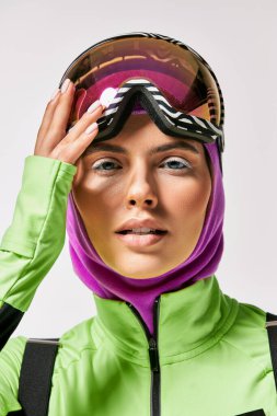 young woman in winter ski clothes with balaclava on head looking at camera on grey backdrop clipart