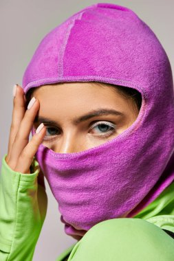 Mor kar maskeli mavi gözlü bir kadının portresi. Gri arka planda eli yüzüne yakın poz veriyor.