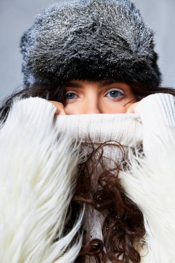Beyaz, sahte kürk ceketli, kışlık şapkalı ve süveterli, gri arka planda poz veren çekici bir kadın, mavi gözlü.