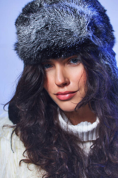 winter beauty, young woman with blue eyes posing in hat with snow on  grey backdrop, cool air
