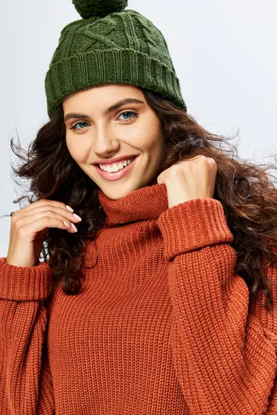 stock image cheerful woman in hat with pom pom and knitted terracotta sweater posing on grey backdrop