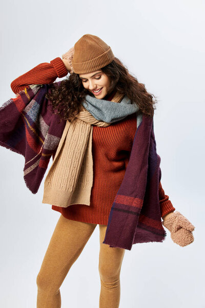 winter style, cheerful young woman in layered clothing, knitted hat and mittens on grey backdrop