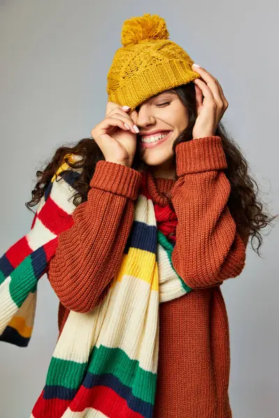 stock image cheerful woman wearing warm bobble hat and sweater with stripped scarf posing on grey background
