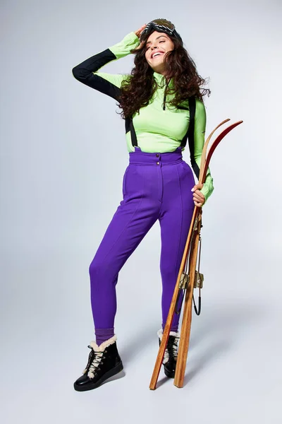 stock image cheerful young woman with curly hair posing in stylish sporty clothes and holding skis on grey