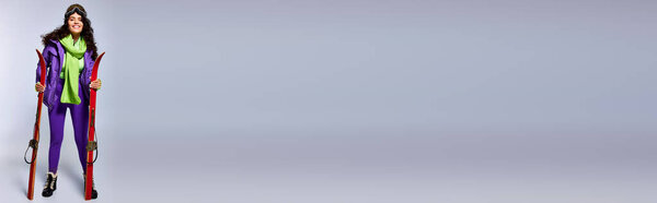 зимнее знамя, положительная женщина с вьющимися волосами, позирующая в активной одежде с пиджаком и лыжами