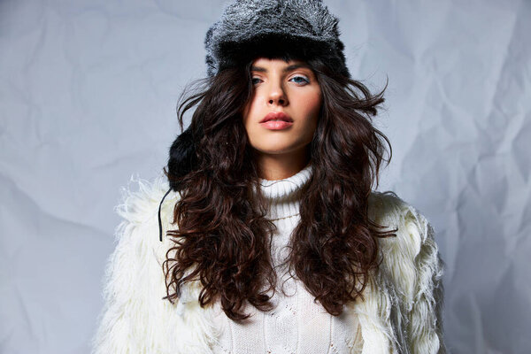 curly woman in faux fur hat and white sweater looking at camera on grey backdrop, winter beauty