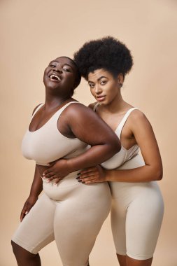 Neşeli ve iri yarı Afro-Amerikalı iç çamaşırlı kadınlar bej renkli kameraya bakıyor, vücut pozitifliği