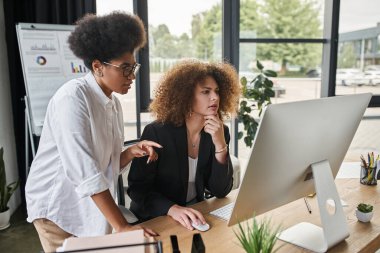 african american businesswoman pointing at computer monitor near colleague working in modern office clipart