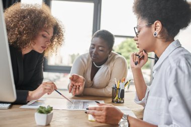 Çok ırklı iş kadınlarından oluşan çeşitli bir grup modern ofiste dokümanların yanında grafiklerle konuşuyorlar.