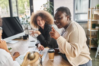 creative team of ambitious multiethnic businesswomen talking near computer and documents in office clipart