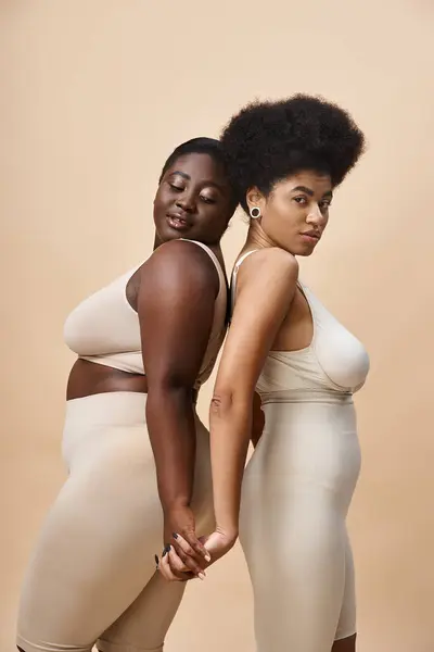 sensual body positive african american women standing back to back on beige, plus size elegance