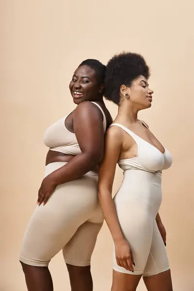sensual body positive african american women standing back to back on beige, plus size elegance