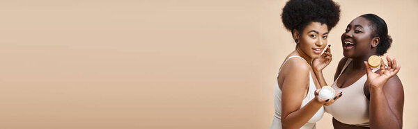 Joyful african american plus size women in lingerie posing with cosmetic cream on beige, banner