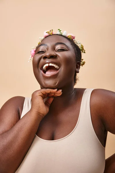 excited plus size african american woman with flowers in hair laughing on beige, body positivity