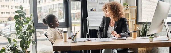 Stock image interracial businesswoman talking near computer monitor and documents at workplace in office, banner