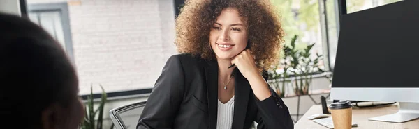 young businesswoman smiling near computer monitor and african american colleague in office, banner