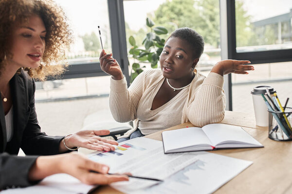 curvy african american businesswoman talking to colleague near documents with graphs in office