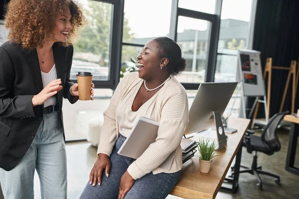 cheerful businesswoman with coffee to go talking to laughing african american colleague in office