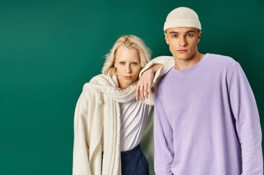 Bereli uzun boylu, yakışıklı bir adam. Sarışın bir kadınla kameraya bakıyor. Turkuazlı.