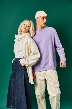 Sarışın kadın, elleri ceplerinde poz veriyor. Bereli uzun bir adamın yanında. Kışın turkuaz giyinmiş.