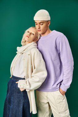 Güzel bir kadın elleri ceplerinde poz veriyor. Bereli uzun bir adamın yanında. Kışın turkuaz giyinmiş.